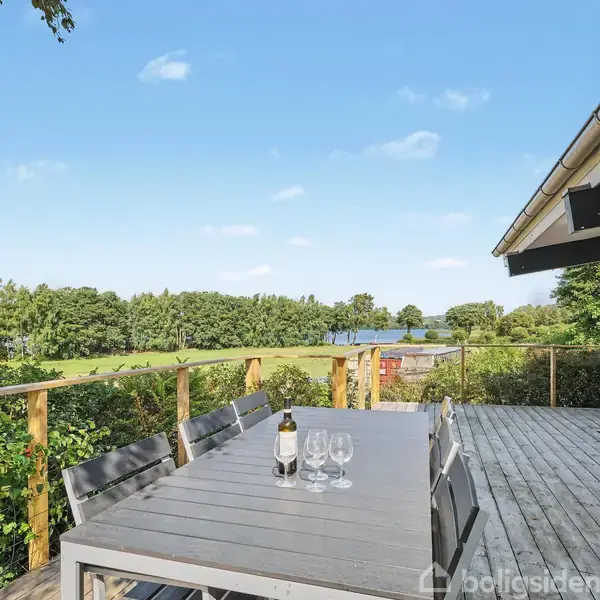 En terrasse med et spisebord og stole står ud mod en udsigt af en grøn have og en sø. Der er en flaske vin og tre glas på bordet.