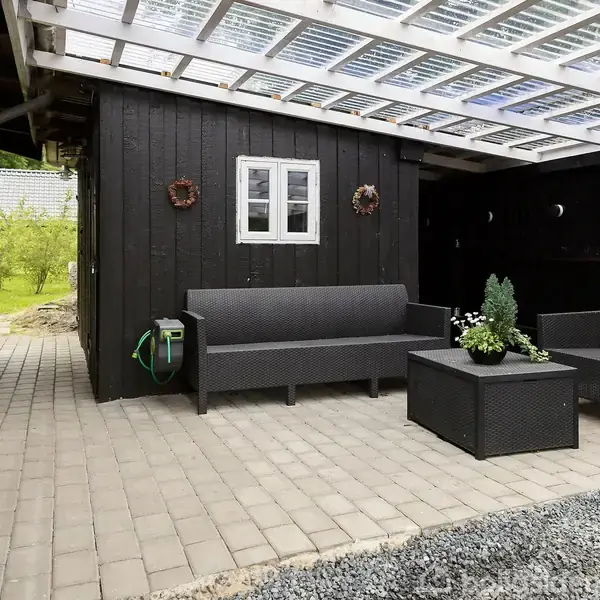 En overdækket terrasse med to sorte sofaer og et kvadratisk bord står på en flisebelagt grund ved et sort træhus. En lille have og stigrus er i baggrunden.