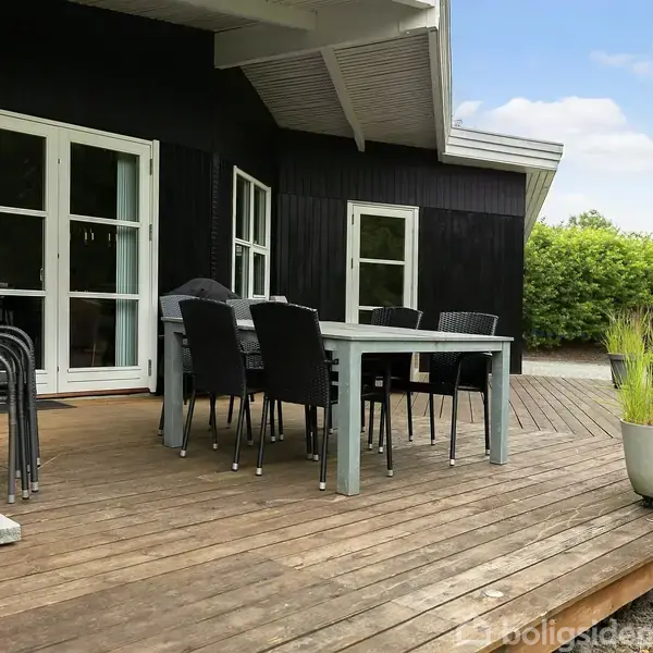 Terrassebord med stole står på en træterrasse foran et sort hus med hvide døre. Terrassen er omgivet af grønne planter og græsplæne, hvilket skaber en indbydende udendørs atmosfære.