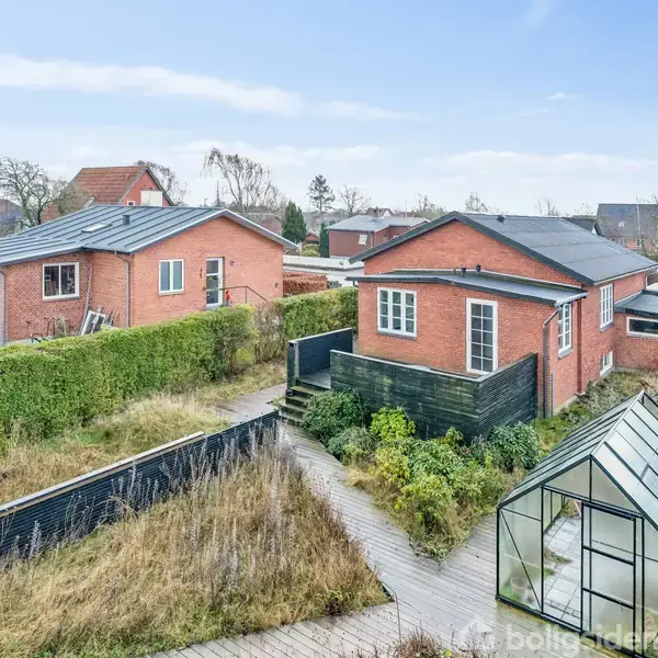 To murstenshuse med røde teglstenstag står i en have med en glaskonstruktion omgivet af græs og buske, beliggende i et forstadskvarter med flere huse i baggrunden.