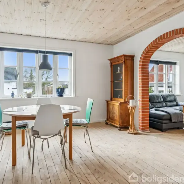 Et spisebord med stole står under en hængelampe i en lys stue med trægulv. En murstensbue fører til en opholdsdel med sofaer. Vinduer slipper naturligt lys ind.