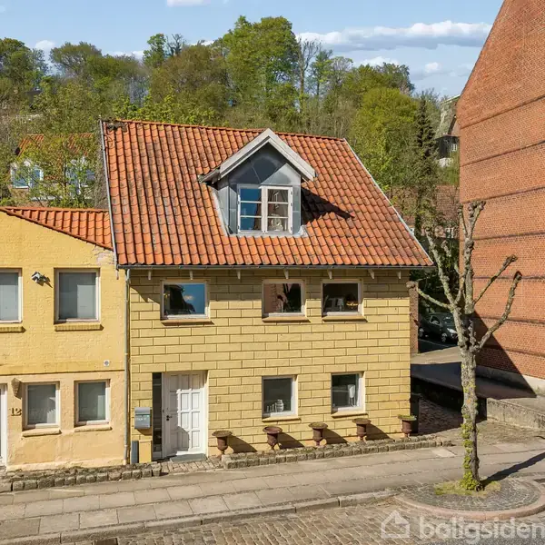 To individuelle to-etagers huse står side om side, omgivet af en brolagt vej. Det ene har et rødt tegltag og en lysegul facade. Baggrunden består af træer og en blå himmel.