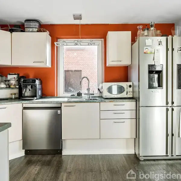 Køkken med grå opvaskemaskine, mikrobølgeovn, og stort køleskab. Skabe omgiver et vindue med røde vægge. På køleskabet hænger magneter og papirer. Trægulve og hvid loftsbelysning.