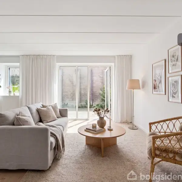 Stue med hvid sofa og rattanlænestol står på et lyst tæppe, omgivet af vægkunst. Et rundt sofabord har en vase med blomster. Store vinduer giver udsigt til en terrasse. Tekst: "Visualisering".