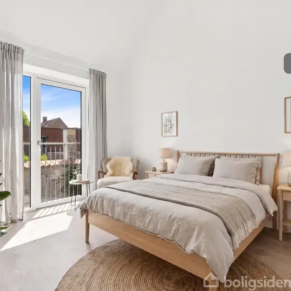 Soveværelse med en dobbeltseng centralt placeret, omgivet af to natborde med lamper. En stor glasdør åbner ud til en balkon, med planter ved siden af døren. Tekst: "Visualisering".
