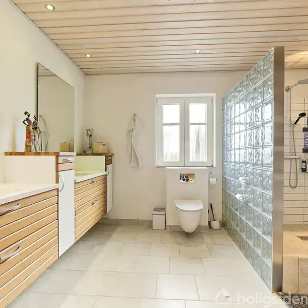 Badeværelse med toilet og bruser. Vasken er på en træbeklædt skabsenhed til venstre. Brusenichen har glasvæg og hylder med produkter. Vindue i midten giver naturligt lys.