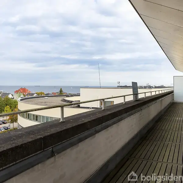 En lang altan med trægulv strækker sig langs en bygning. Den har udsigt over hustage, træer og havet under en delvist overskyet himmel.