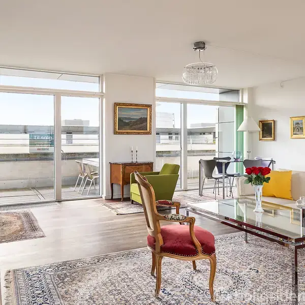 Stue med elegante møbler står stilfuldt arrangeret. En glasbord med en buket roser hviler foran en sofa. Store vinduer giver udsigt til en balkon.