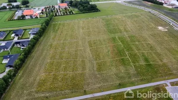 En stor, åben mark med tydelige græsstier set fra luften. Ved siden af ligger adskillige moderne huse og en landevej, omgivet af grønt landskab.