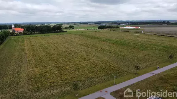 En stor græsmark strækker sig ud i landskabet, omkranset af en bred sti med få træer. I baggrunden ses en kirke med rødt tag og flere bygninger.
