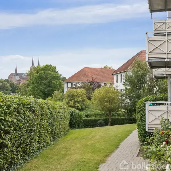 En sti løber langs en hæk, der omgiver boliger med altaner. I baggrunden ses træer og et par kirketårne mod en blå himmel.