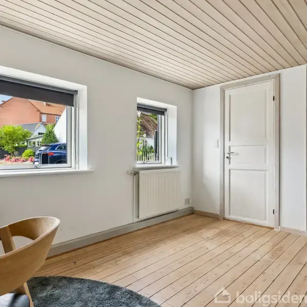 Et lyst rum med trægulv og træloft. To små vinduer ved siden af en radiator. Til venstre ses et rundt spisebord med stole, og til højre en sofa.