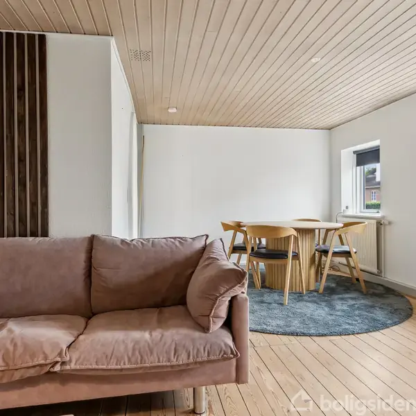 En brun sofa står i en lys stue, ved siden af et spisebord med fire stole på et rundt tæppe. Der er hvide vægge og trægulv med store vinduer, der slipper lys ind.