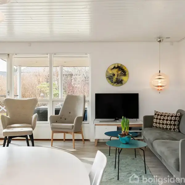 En stue med grå sofa og to lænestole omkring et lille bord. Et TV står på en hvid reol. I forgrunden ses et spisebord med gule blomster. Et vindue viser en have udenfor.