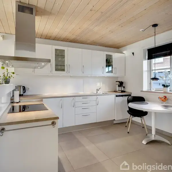 Et moderne køkken er indrettet med hvide skabe, en ovn og komfur. En emhætte hænger over komfuret. Ved vinduet står et rundt bord med to stole.