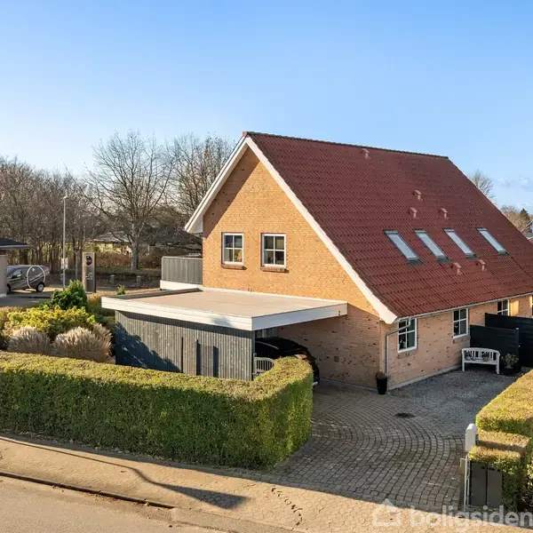 Et to-etagers hus med rødt tag og flere ovenlysvinduer, stående på en grund omkranset af hække og en flisebelagt gårdsplads, beliggende i et boligområde med træer i baggrunden.