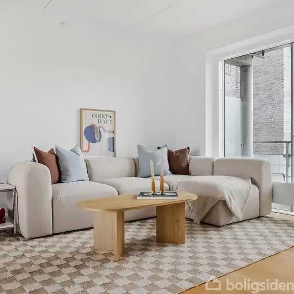 En lys stue har en beige sofa med puder, et træbord med stearinlys, og et mønstret tæppe. Væggene er hvide, en plakat hænger, og store vinduer viser murværk udenfor.