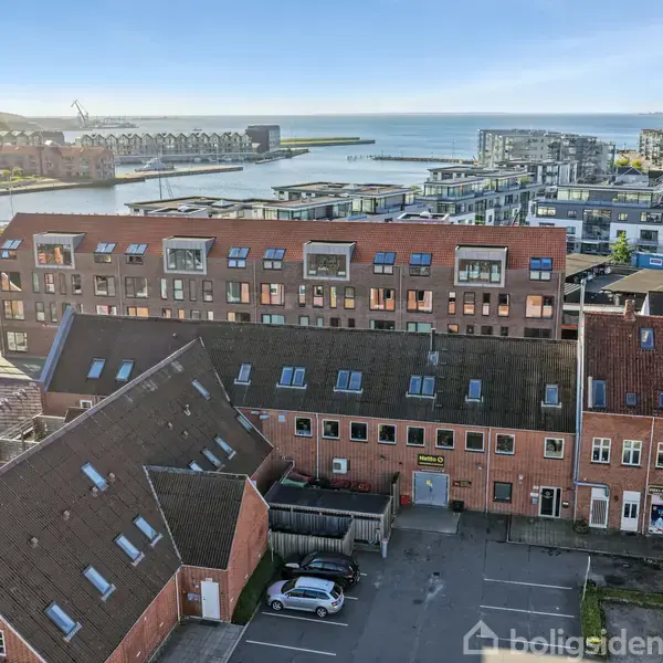 Rødt murstensbyggeri med skråt tag står stille i solrigt havneområde; omkringliggende bygninger og vand skaber en rolig, urban atmosfære.