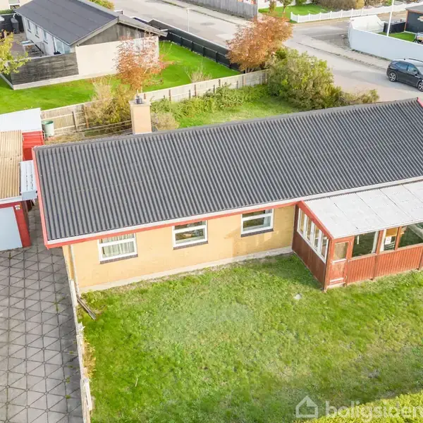Hus med fladt tag og rød garage, omgivet af en grøn have. Et drivhus er ved siden af huset. Beliggende i et beboelsesområde med huse og biler på vejen.