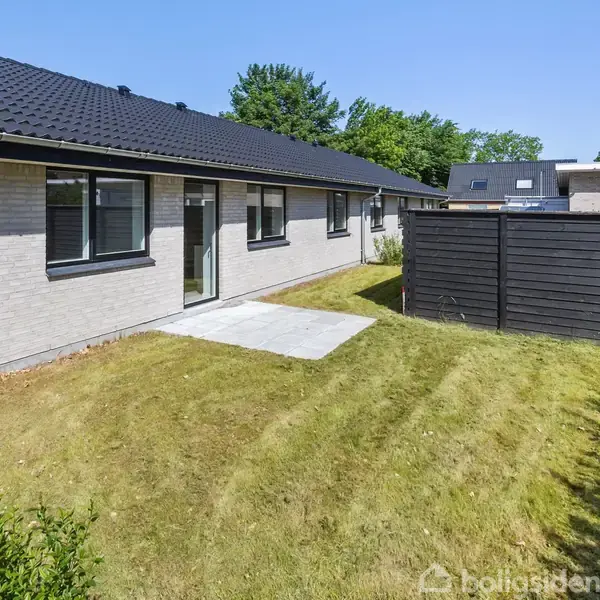 En lav, murstensbeklædt bygning med sorte taget, beliggende i en have med en lille flisebelagt terrasse og en træhegn på en solrig dag.