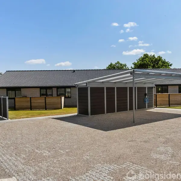 Carport står stille på en grusbelagt parkeringsplads foran rækkehus, omkranset af græsplæner og få træer under en blå himmel med få skyer.