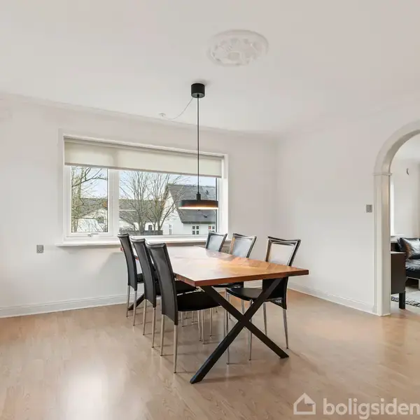 Et spisebord med otte stole står centralt i et lyst rum med trægulv. Ved siden af er der en stueadgang gennem en bue, og udenfor ses træer.