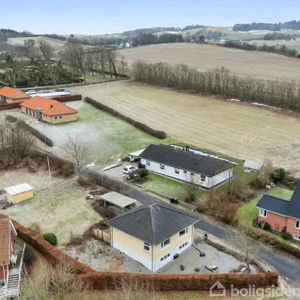 Flere huse ligger ved en vej i et landligt område omgivet af åbne marker og træer. I baggrunden ses en kirke. Boligerne har hække og små haver.