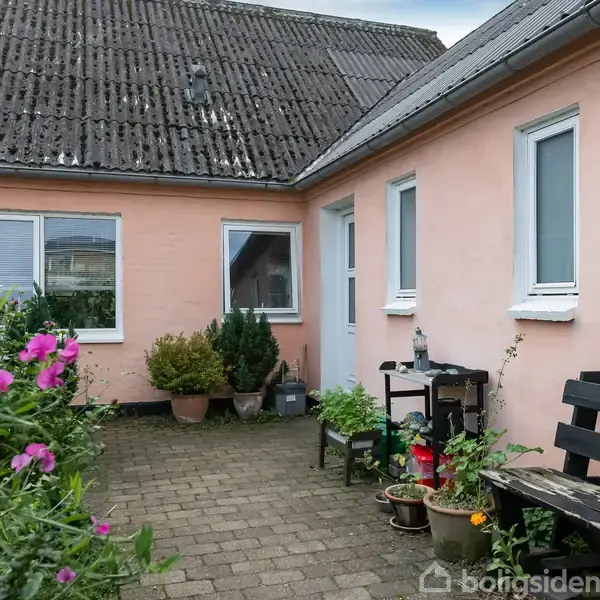 Et lyserødt hus med sort tag danner en gårdhave med fliser, hvor planter og blomster i krukker er placeret. Der står en bænk og et lille bord ved husmuren.