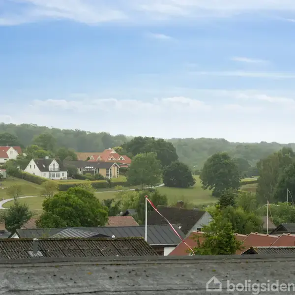Hustage ses i forgrunden, mens en gruppe hvide og røde huse ligger oven på bakkede, grønne områder i baggrunden under en blå himmel.