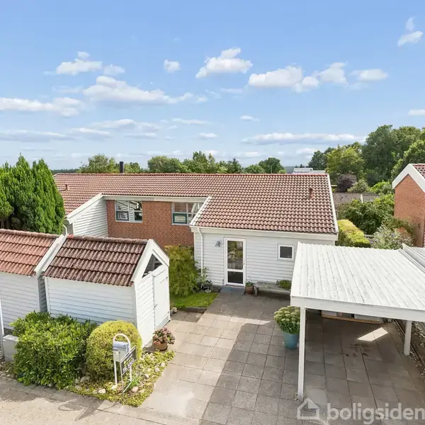 Et hvidt hus med rødt tag står stille. Til venstre ses en carport og en tilhørende garagebygning. Omgivet af en velplejet have og omgivelser med træer.