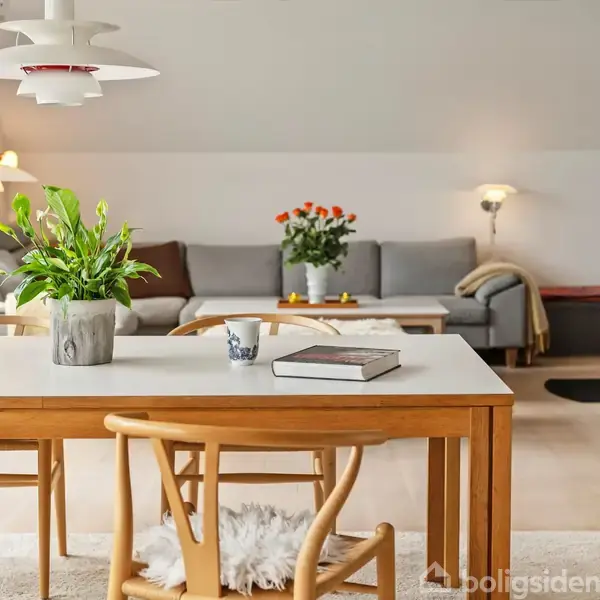 Et spisebord med en grøn plante og en bog står i midten af en lys stue. I baggrunden er en grå sofa og et sofabord med blomster. En lampe hænger over bordet.
