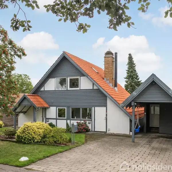 Huset, med et moderne design, har rødt tegltag og grå facade. Det står i en velplejet have omgivet af træer og buske, med indkørsel og carport ved siden af.