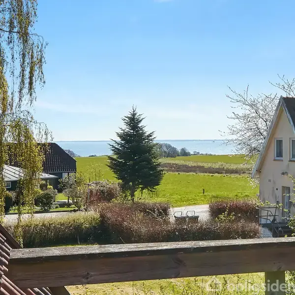 Huse med skrå tage står i en landlig omgivelser med udsigt over en græsmark og træer. I baggrunden ses havet under en klar blå himmel.
