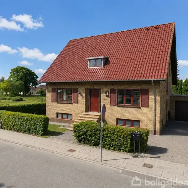 Et murstenshus med rødt tag ligger ved en vej. Huset har en dør på forsiden og en garage ved siden. Haven er velplejet med hække omkring. Himmel med få skyer.