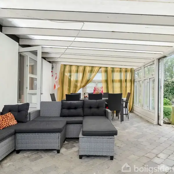 Grå sofa står i en lys udestue med hvide bjælker, der fører ud til en have. Gardiner trukket til siden afslører et spisebord med stole nær døren.