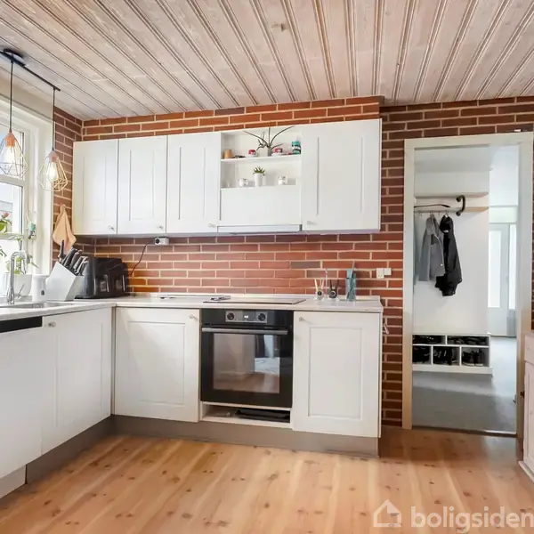 Køkken med hvide skabe og sort ovn integreret i en murstensvæg, træloft og gulv, og planter i vinduet. Mørkt tøj hænger i gangen til højre.