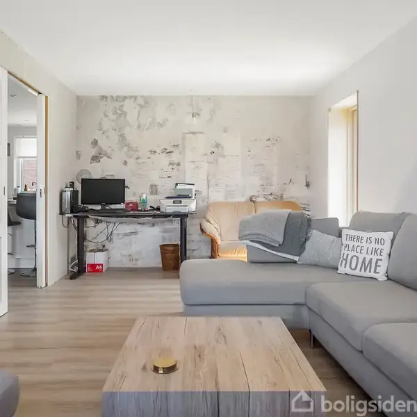 En stue med en grå hjørnesofa, hvor en pude har teksten "There is no place like home." En træbord står foran sofaen. Bagved ses en arbejdsstation med computer.
