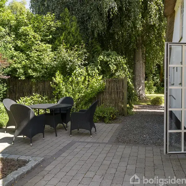 Et terrasseområde med sorte havemøbler står på en flisebelagt overflade ved siden af en hvid bygning. Omgivet af tætte grønne buske og træer i baggrunden.