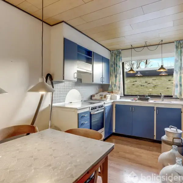 Et køkken med et spisebord og fire stole, hvor en lampe hænger over bordet. Blå og hvide køkkenelementer findes til venstre og højre. I baggrunden ses en dør til et andet rum.