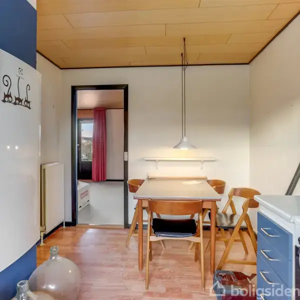 Et køkkenbord med fire træstole står under en hængende lampe. Køkkenet har hvide og blå skabe. En dør til et rum med røde gardiner er synlig i baggrunden.