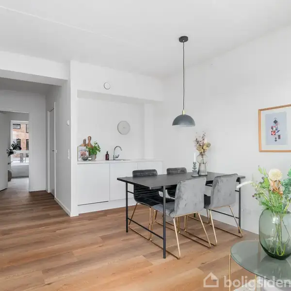 Spisebord med fire stole under en hængende lampe i et lyst, moderne køkken-alrum med trægulv. Væggene er dekoreret med to indrammede billeder.