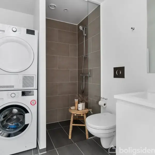 Stabel af vaskemaskine og tørretumbler står i et moderne badeværelse med brusekabine, toilet og vask. En træskammel med toiletartikler står ved siden af.