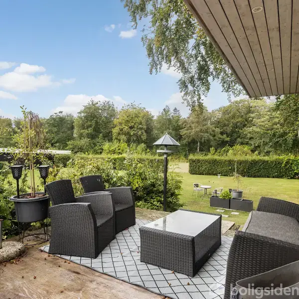Havemøbler står arrangeret på en terrasse med fliser; sofa og stole omkring et lavt bord. I baggrunden ses en velplejet have med buske og træer.