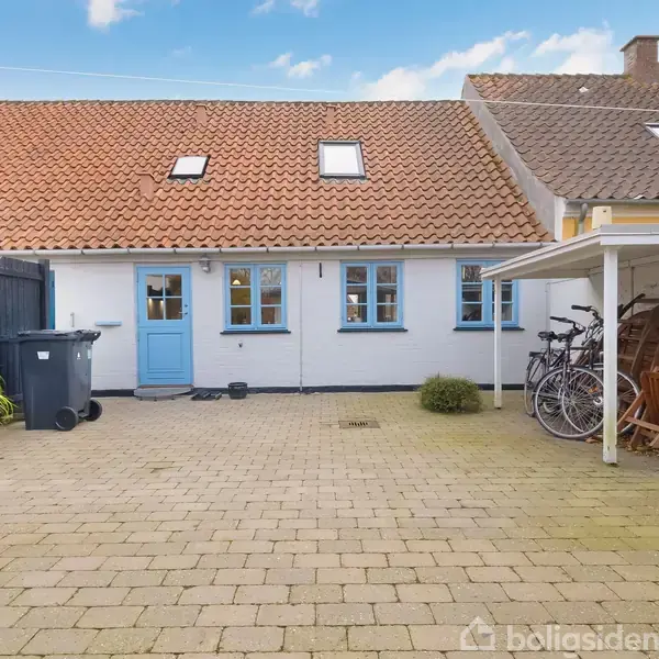Et hus med blå dør og vinduer står i en brostensbelagt gårdhave. Cykler står under et halvtag til højre. En skraldespand og planter er til venstre.