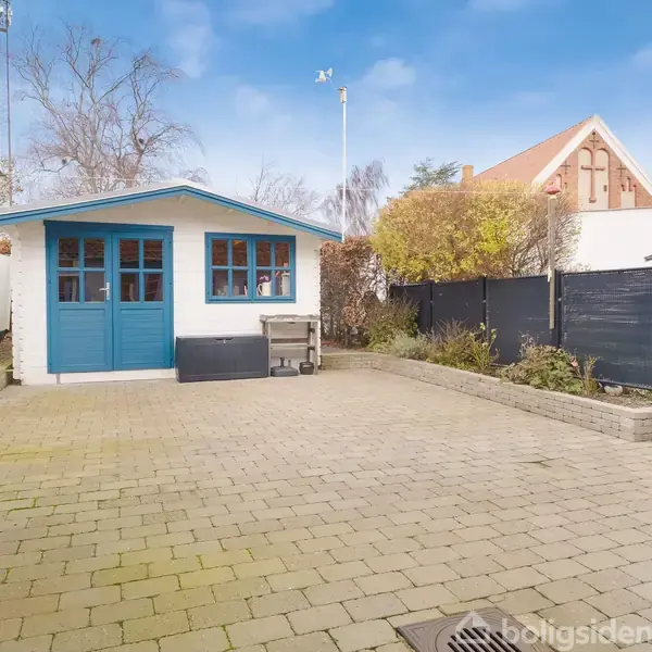 Et lille skur med blå døre står for enden af en flisebelagt gårdhave. Skuret er omgivet af planter og høje hegn, alt under en blå himmel.