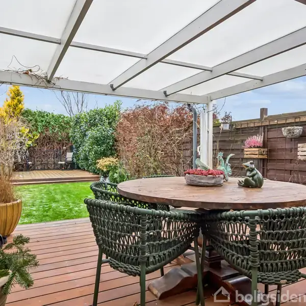 En rund træbord med flettede stole står på en overdækket terrasse. I baggrunden ses en have med grøn græsplæne og omgivet af buske og hegn.