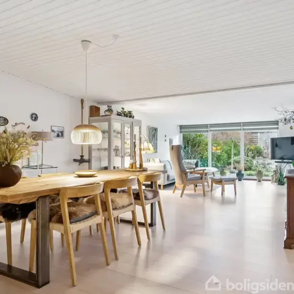 Spisebord med træstole dekoreret med vase og skål, placeret i rummelig stue. Store vinduer lyser rummet op, som også indeholder reoler og sofa.