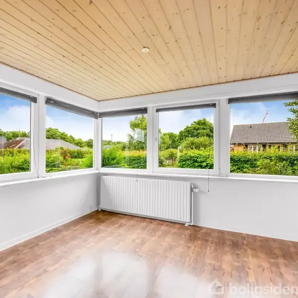 Et tomt rum har trægulv og lyse træpaneler i loftet. Store vinduer giver udsigt til en grøn have og omkringliggende huse. En radiator er monteret under vinduerne.