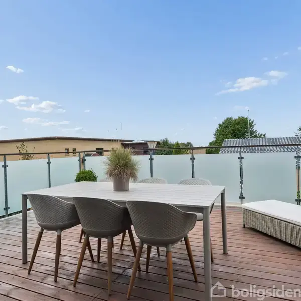 En udendørs terrasse med et hvidt bord, fire stole og en liggestol. Bordet har en plante i midten. Terrassen har glasrækværk og udsigt til nabobygninger og træer.