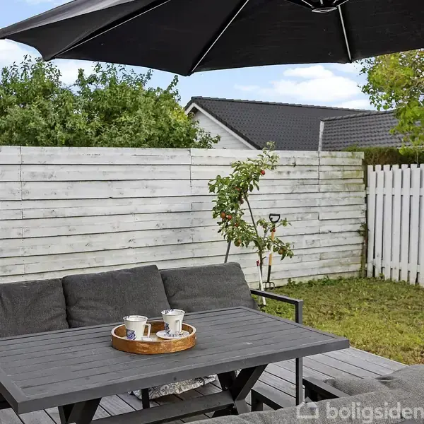En terrasse med et bord, hvorpå der står et fad med kopper. I baggrunden er der et lille træ med frugt ved et hvidt plankeværk i en have.